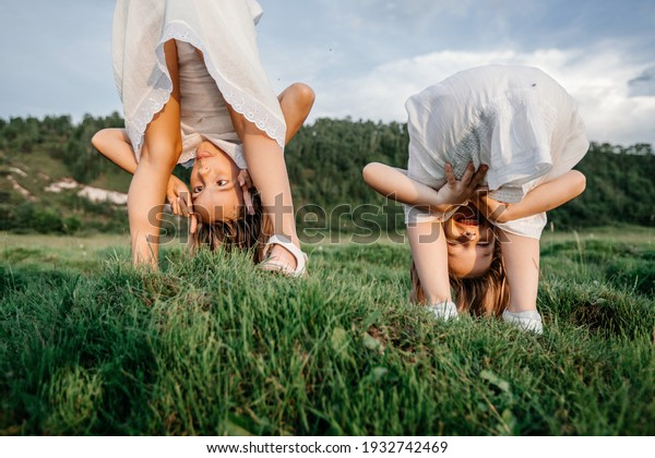 Upside down photo of happy children in white\
dresses play on the grass and smile in summer. Funny portrait of\
two sisters. Children\'s\
friendship