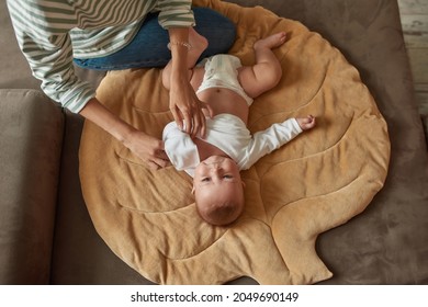 Upside Down Portrait Of Cute Satisfied Baby, Lying Down On Large Flat Pillow, Enjoying Morning Dressing Routine. Unrecognisable Woman Taking Off White Bodysuit From Her Child. Top View.