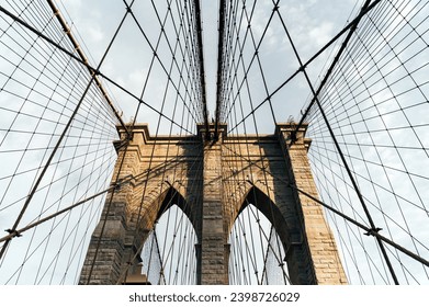 Upward angle view of the Brooklyn Bridge's iconic cables and stone pillars against a cloudy sky in Manhattan, New York - Powered by Shutterstock