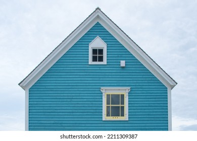 An upward view of a cloudy sky and an exterior gable end of a blue wooden house with two small windows.The windows have yellow and white trim. The peaked roof of the building has white decorative trim