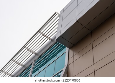 The upward view of a modern commercial building with beige and grey metal composite panels, blue glass windows, and a slated grate on the edge of the roof. The sky is cloudy and white in color. - Powered by Shutterstock