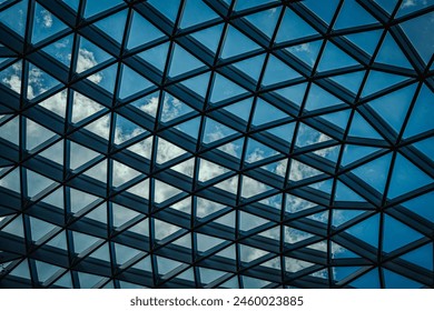 An upward view of a modern geometrical glass ceiling, showcasing a complex network of triangular panels framing a blue sky with fluffy clouds. - Powered by Shutterstock