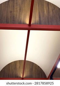 Upward view of a modern interior ceiling featuring red structural beams, curved white sections, and decorative wood-finish panels. Contemporary architectural detail suitable for interior design, construction, and commercial building concepts