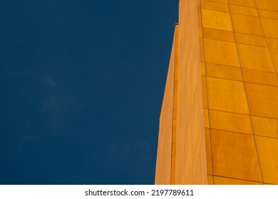 The Upward View Of Pale Wood Grain Composite Board Panels On The Exterior Of A Tall Modern Building. There's A Row Of Glass Windows Between The Two Sections And A Deep Blue Sky In The Background.