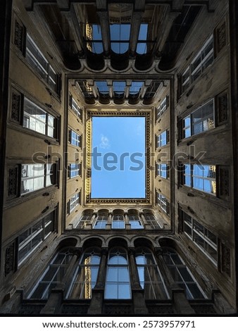 An upward view of a rectangular courtyard surrounded by a historic building, framing the clear blue sky. The architecture features symmetrical windows and ornate details, creating a striking geometric