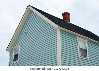 An upward view of a vintage blue wooden building with thick white trim, a small double hung window, with four panes of glass and a frame shingled roof. The roof has white trim and black shingles.