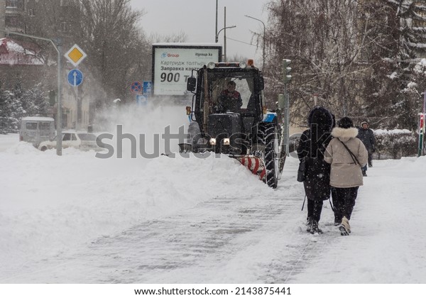 Uralsk,\
Kazakhstan (Qazaqstan), 11.03.2022 - Snow removal by a tractor in\
the city of Uralsk, a tractor with a bucket and a brush for snow\
removal, a tractor removes snow on the\
sidewalk