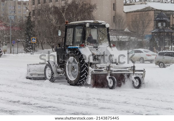 Uralsk,\
Kazakhstan (Qazaqstan), 11.03.2022 - Snow removal by a tractor in\
the city of Uralsk, a tractor with a bucket and a brush for snow\
removal, a tractor removes snow on the\
sidewalk