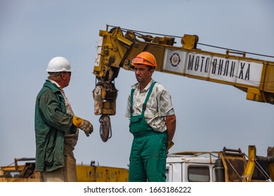 Uralsk Region, Kazakhstan - May 24 2012: Oil Deposit Zhaikmunai. Oil Drilling Rig. Two Workers Talking. Yellow Crane, Crane Hook With Steel Cable. Blue Sky.