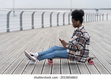 Urban african american girl sitting on longboard with smartphone in hand. Black female skater female texting outdoors in park using 5g connection. Skateboarder concentrated on message in mobile phone