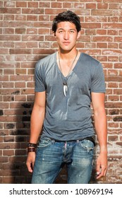 Urban Asian Man. Good Looking. Cool Guy. Wearing Grey Shirt And Jeans. Standing In Front Of Brick Wall.