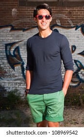 Urban Asian Man With Red Sunglasses. Good Looking. Cool Guy. Wearing Dark Blue Shirt And Green Shorts. Standing In Front Of Brick Wall With Graffiti.