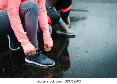 Urban athletes lacing sport footwear for running over asphalt under the rain. Two women getting ready for outdoor training and fitness exercising on cold winter weather.