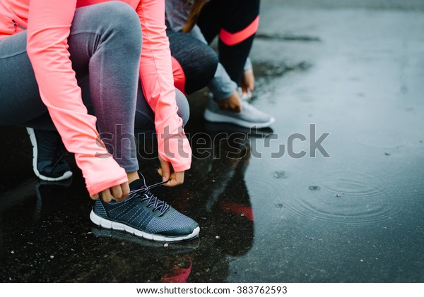 Urban athletes lacing\
sport footwear for running over asphalt under the rain. Two women\
getting ready for outdoor training and fitness exercising on cold\
winter weather.