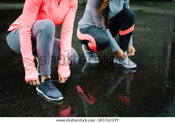 Urban athletes lacing\
sport footwear for running over asphalt under the rain. Two women\
getting ready for outdoor training and fitness exercising on cold\
winter weather.