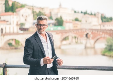 Urban Businessman Wearing Jacket And Shoulder Bag. Handsome Male Model In His 45s. Young Entrepreneur Walking In The Street And Holding A Mobile Smartphone Using App Text Sms Message