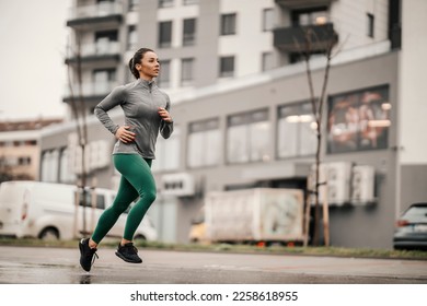 An urban female runner is running on the rainy day on city street.