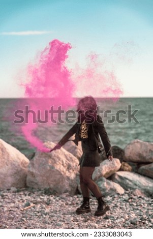 urban girl on the beach playing with a pink smoke flare in the foreground and with big stones and the sea in the background on a day with a completely clear sky.