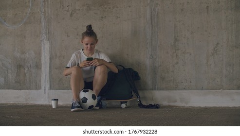 Urban Girl Using Mobile Phone. Caucasian Teenager Football Soccer Player Sitting On Skateboard Texting On Smartphone Inside Empty Covered Parking Garage. 4K UHD Slow Motion RAW Graded Footage