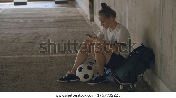 Urban girl using mobile phone. Caucasian teenager\
football soccer player sitting on skateboard texting on smartphone\
inside empty covered parking garage. 4K UHD slow motion RAW graded\
footage