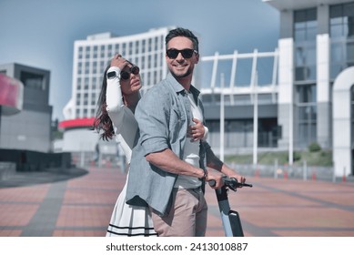 Urban journey. A stylish couple, a young man and a girl, ride together and smile on an e-scooter along a sunny street against the backdrop of modern city architecture - Powered by Shutterstock