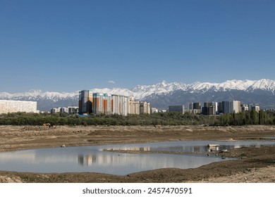 Urban landscape, modern buildings rise against the awe-inspiring backdrop of snow-capped mountains. Clear blue sky reflects off pond water. Unique beauty of a city nestled at foot of towering peaks - Powered by Shutterstock