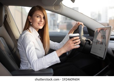 Urban Lifestyle, Vehicles And People Concept. Portrait Of Happy Young Caucasian Business Woman Sitting In Modern Electric Car And Drinking Take Away Coffee, Waiting For Battery Charging