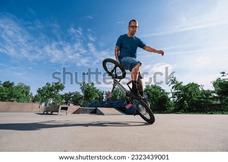 An urban middle-aged skilled bmx rider is practicing freestyle tricks on his bike in a skate park. A mature tattooed master is balancing on one wheel on his bike in a skate park. Stock photo © 