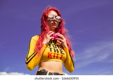 Urban Portrait Of A Girl With Red Hair, A Yellow T-shirt And Ripped Jeans, Pink Headphones, Listens To Music. 2000s Style, 2020 Trends