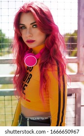 Urban Portrait Of A Girl With Red Hair, A Yellow T-shirt And Ripped Jeans, Pink Headphones, Listens To Music. 2000s Style, 2020 Trends