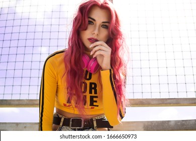 Urban Portrait Of A Girl With Red Hair, A Yellow T-shirt And Ripped Jeans, Pink Headphones, Listens To Music. 2000s Style, 2020 Trends