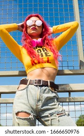 Urban Portrait Of A Girl With Red Hair, A Yellow T-shirt And Ripped Jeans, Pink Headphones, Listens To Music. 2000s Style, 2020 Trends