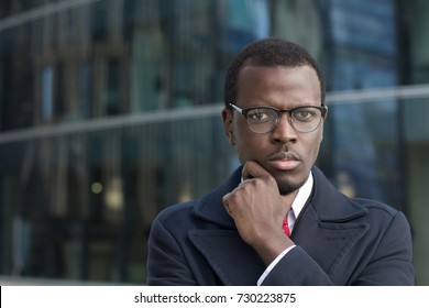 Urban portrait of handsome African businessman dressed in smart coat in city street pressing fist to his chin while thinking hard and trying to make best decision for development of his business