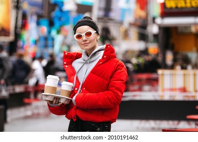 Urban Portrait Of Stylish Young Girl On New York Street With Coffee To Go. Casual Modern Woman Wearing Trendy Sunglasses, Red Jacket And Street Style Clothes Over Time Square, NYC Downtown Background