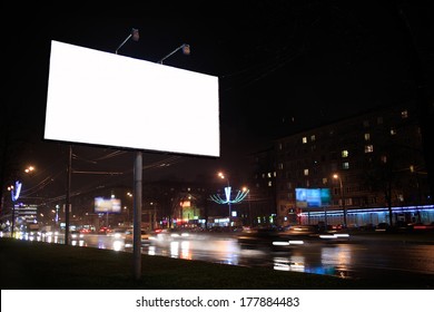 Urban Scene With An Illuminated Empty White Billboard On The Side Of A Street With Cars In Motion And A Block Of Flats In The Background, By Night