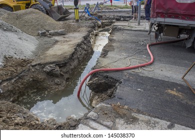 Urban Scene, A Utility Crew Works. An Open Trench Is Flooded, Deep Under The Road Surface Is The Broken Pipe.