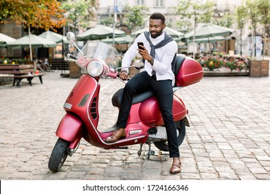 Urban Style, City Business. Young African American Bearded Guy, Wearing Smart Casual Clothes, Using His Mobile Phone, Sitting On The Red Scooter On The Cite Square Outdoors