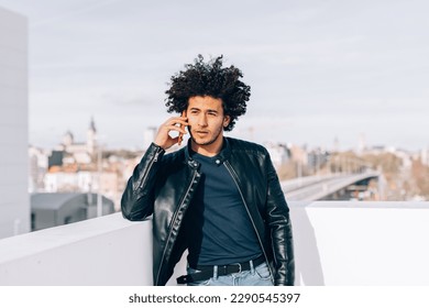 Urban style portrait of a handsome North African guy while he talks on the phone with a captivating look, as he looks into the camera, dressed in a leather jacket