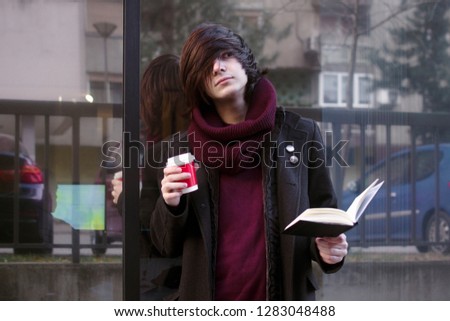 The urban style student boy with book and coffee is standing outdoors at the street and resting.