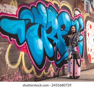 Urban stylish travel: young latina brunette with fashion look and pink suitcase in front of a lively street art mural at downtown - Powered by Shutterstock