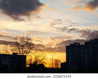 Urban sunset with dramatic clouds over city buildings and bare trees. Warm golden light, silhouetted skyline, evening atmosphere, cityscape at dusk with copy space.