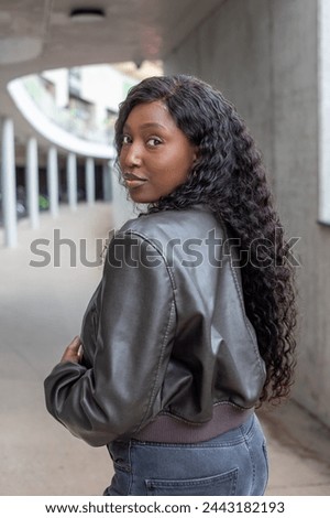 In an urban underpass, a young African woman with long curly hair turns to glance over her shoulder. She exudes casual confidence and style in a sleek leather jacket, against a backdrop of concrete
