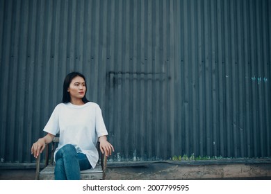 Urban Young Asian Woman Sitting On Chair Near Blue Wall On The Street Outdoors With Copy Space Template