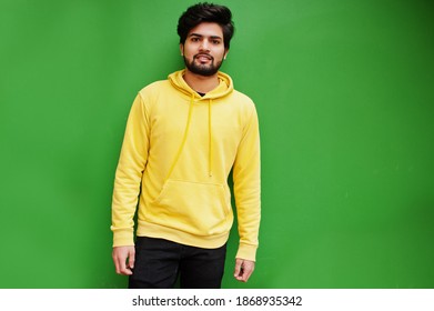 Urban Young Hipster Indian Man In A Fashionable Yellow Sweatshirt. Cool South Asian Guy Wear Hoodie Isolated On Green Background.