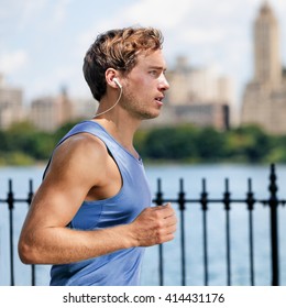 Urban Young Man Running In City Park Listening To Music With Wireless In-ear Earphones Living A Healthy Active Lifestyle. Male Runner Blue Top Working Out Cardio Exercise Workout In Summer.