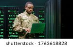 US army male African American officer in uniform stading at server wih secret data and using laptop computer to check the information. Military monitoring service room. Army networking concept.