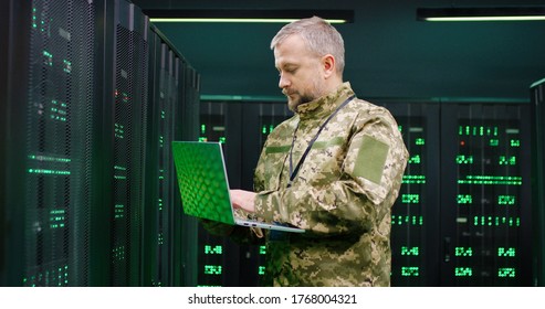 US Army Senior Caucasian Male Soldier Working At Laptop Computer In Dark Room With Monitors. Technician From Military Forces Typing On Keyboard And Sending Secret Data Of Surveillance.