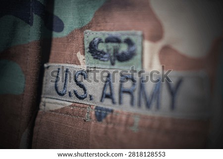 U.S. Army uniform patch close-up, bold letters on light fabric, smaller insignia above, muted earth tones, soft lighting, textured material, indoor display setup