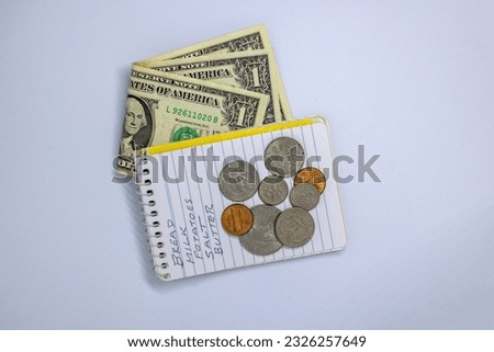 US dollar banknotes folded under a notebook, and coins over a handwritten shopping list isolated on white background