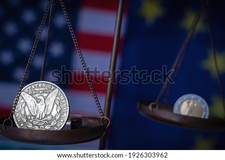 US Dollar coin and EU currencies on weighing scale against Unated States and European Union flags as symbol of  trade war, economic conflict and  relations. Selective focus on Dollar.